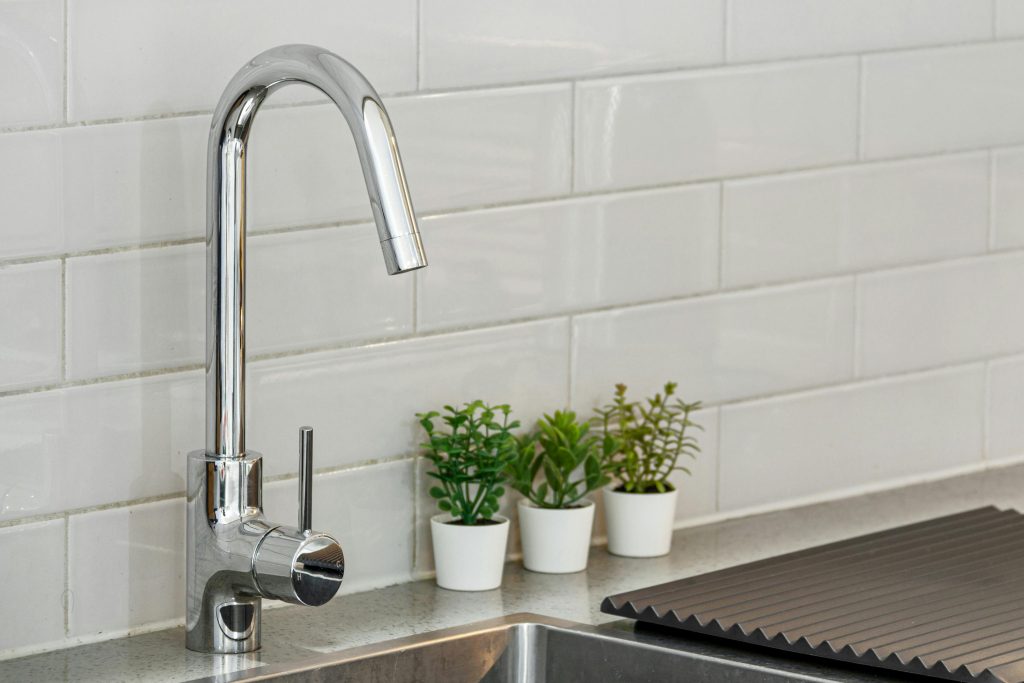 A sleek kitchen sink with chrome faucet and potted plants on a tiled backsplash.